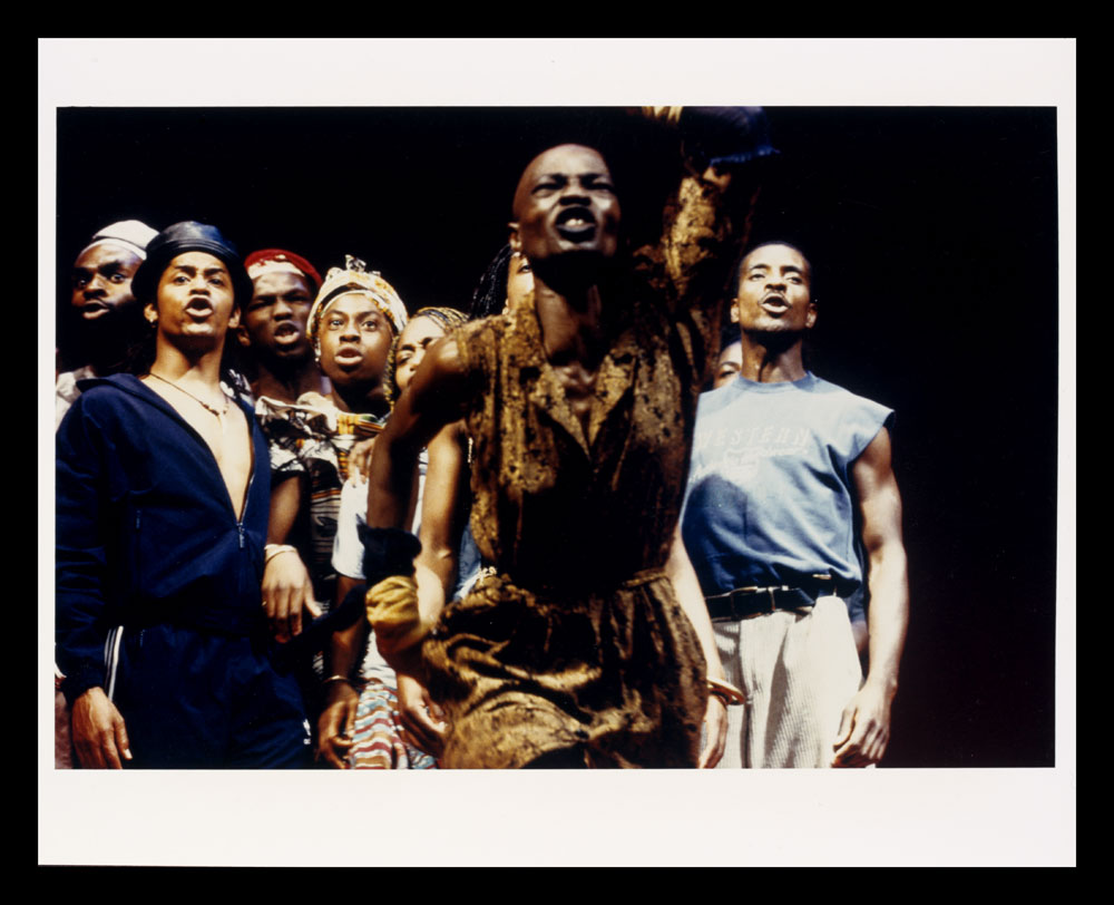 Colour photo of actors on stage, woman in yellow dress in front, from Zumbi